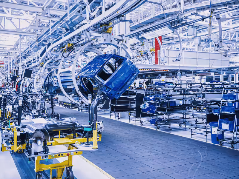 Alt text: A single blue car chassis is being rotated by a large white robotic arm in an automated car factory.