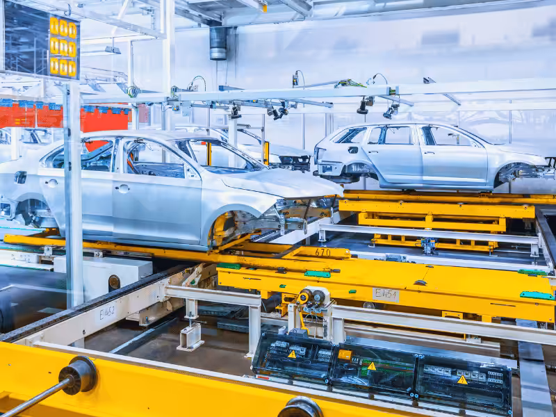 Two unpainted silver car bodies are moving on yellow tracks through an automated car factory, guided by overhead robot systems.